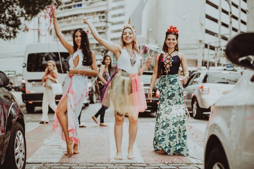 Young cheerful barefoot women in stylish apparel with raised arms standing on street pavement while looking at camera