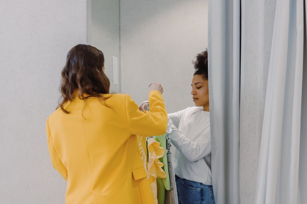 A personal stylist helps a client with clothing choices in a fitting room, focusing on fashion styling.